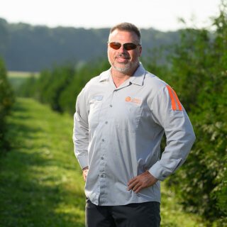 Ken Corrin, Proprietor of Rolling Fields Tree Farm and GreenGiantTrees.com, standing proudly beside a row of trees with hands on hips.